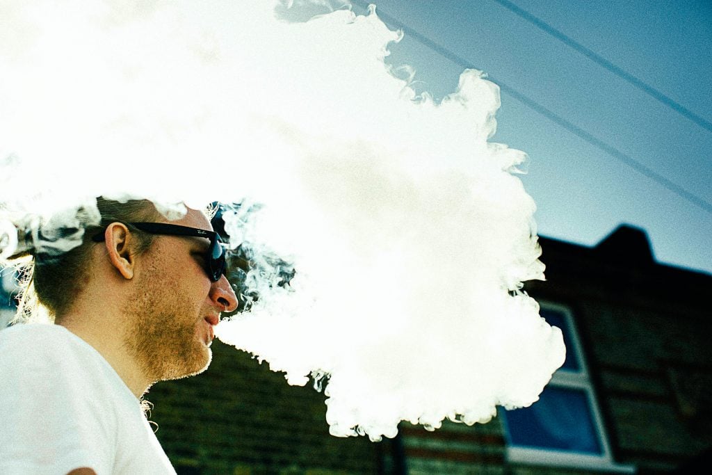 man blowing thick white smoke on air overlooking black building wall at daytime